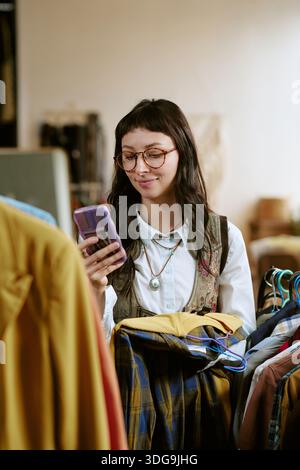 Jeune femme biraciale comparant des vêtements de seconde main à l'aide d'un smartphone Banque D'Images