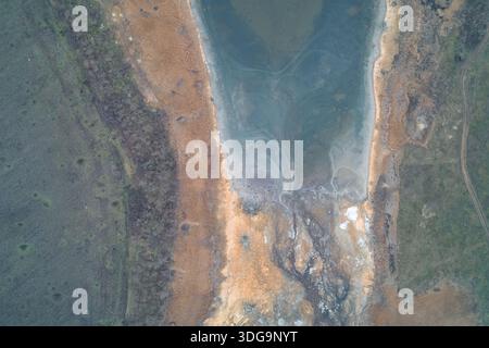 Une vue aérienne complexe d'un rivage d'un lac en recul révélant des couches de sédiments, des roseaux bruns et de l'eau stagnante. Le paysage texturé met en évidence les changements hydrologiques et la beauté brute d’un écosystème de zones humides asséchées. Banque D'Images