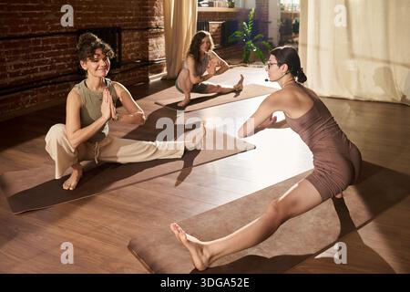 Trois jeunes femmes adultes pratiquant des étirements de yoga sur des tapis en studio, instructeur dirigeant une séance de groupe, tous axés sur des exercices de flexibilité Banque D'Images