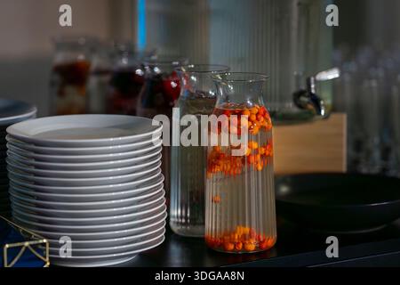 Carafes en verre remplies d'eau infusée et de baies d'argousier sur une table de buffet moderne avec des assiettes blanches empilées. Banque D'Images