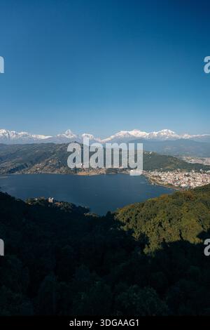 Vue sur un lac serein niché au milieu de collines verdoyantes, avec des montagnes enneigées scintillant sous un vaste ciel bleu, Himalaya, Annapurna, Népal. Banque D'Images