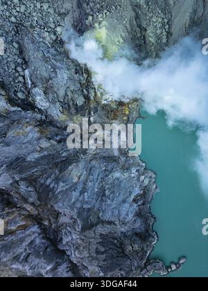 Vue aérienne du lac turquoise rencontrant le paysage texturé et rocheux, avec des panaches de fumée blanche s'élevant dans l'atmosphère, Banyuwangi, Java oriental, Banque D'Images