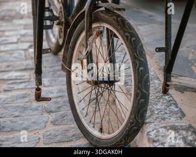 Roue de vélo rouillée et détail de cadre garé par un trottoir sur un trottoir pavé. Banque D'Images