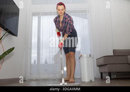 Une femme portant une chemise à carreaux, un bandeau et des gants rouges nettoie le sol de son salon à l'aide d'une serpillière. Elle est pieds nus et concentrée sur sa corvée près d'une fenêtre et d'un canapé. Banque D'Images