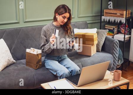 Assise confortablement sur un canapé, la femme montre sa carte de crédit tout en utilisant son smartphone pour faire des achats en ligne. Banque D'Images
