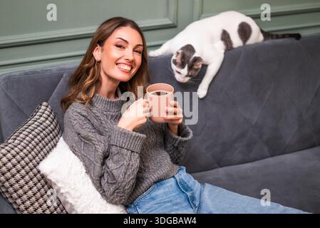 Femme adulte heureuse profite du temps de détente à la maison. Elle est assise au canapé et boit du café, tandis que le chat est allongé à côté d'elle. Banque D'Images