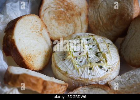 Camembert cuit au four aux herbes et à l'huile d'olive, servi avec des tranches de pain grillées sur du papier sulfurisé. Apéritif maison confortable photographié en naturel Banque D'Images