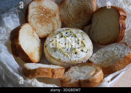 Camembert chaud garni d'herbes et d'huile d'olive, entouré de tranches de pain grillées. Cuisine réconfortante maison et hors-d'œuvre de partage Banque D'Images
