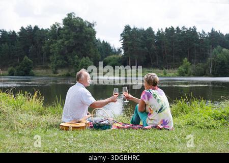 charmant couple de personnes âgées au lac ayant un pique-nique, faisant un toast sur la rive du lac un jour d'été. Staycations, voyage hyper-local, sortie en famille, escapade. || autorisation du modèle Banque D'Images