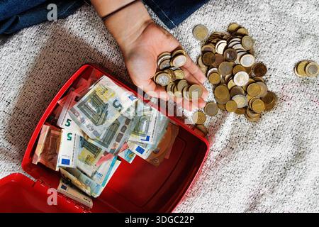 Vue de dessus de la main d'une jeune femme tenant des pièces en euros à côté d'une caisse ouverte avec des billets en euros sur une couverture de lit Banque D'Images