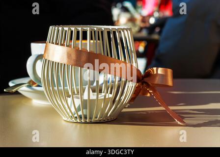 Plateau de café ensoleillé avec un bougeoir décoratif enveloppé d'un noeud en ruban, avec une tasse à café et une soucoupe légèrement floues en arrière-plan. Banque D'Images