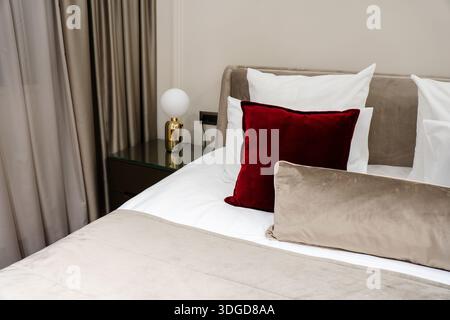 Lit d'hôtel moderne avec draps blancs, tête de lit beige et oreillers décoratifs en velours rouge dans l'élégant intérieur de la chambre Banque D'Images