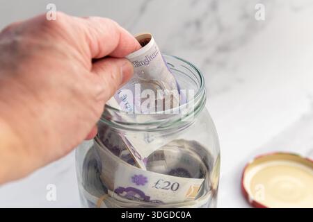 Un gros plan d'une personne économisant de l'argent en mettant des billets de banque Sterling Pound dans un bocal en verre. Banque D'Images