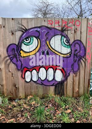 Visage coloré de graffiti de dessin animé sur une clôture en bois, avec des yeux larges et un sourire dentelé, des contours noirs audacieux et un style Street-art. Banque D'Images