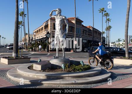 Statue de surfeur se dresse à une intersection de rue près de la jetée de Newport Beach alors qu'un cycliste passe par, Newport Beach, Californie. Banque D'Images