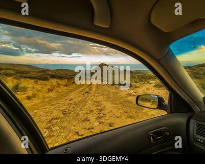 La vue panoramique de l'intérieur de la voiture capture le chemin de terre menant au lac. Les montagnes et le ciel nuageux améliorent l'humeur aventureuse pendant le coucher du soleil. Banque D'Images