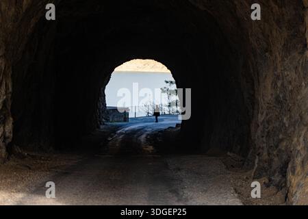 Vue de l'intérieur d'un tunnel rocheux sombre regardant vers un lac bleu vif. Une silhouette d'une personne se dresse à la sortie du tunnel. Contraste spectaculaire et Banque D'Images