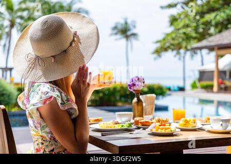 Femme en chapeau de soleil profite du petit déjeuner tropical près de la station balnéaire au bord de la piscine, tenue Banque D'Images