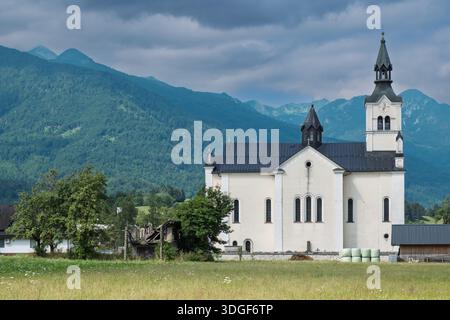 Église de Nicholas à Bohinjska Bistrica, Slovénie, situé dans un champ avec des balles de foin et des montagnes sous un ciel nuageux. Banque D'Images