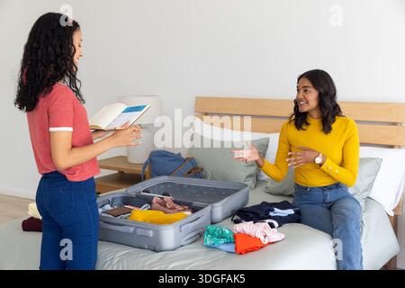 Maman diversifiée et fille adolescente emballant la valise dans la chambre à coucher en utilisant le guide de voyage et les vêtements Banque D'Images