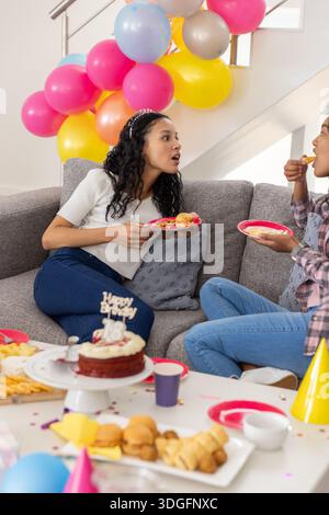 Maman diversifiée et fille adolescente célébrant sur le canapé dans le salon avec gâteau Banque D'Images