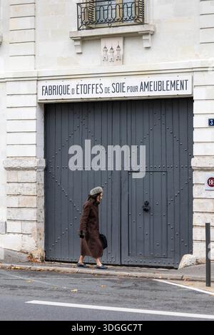 Femme dans un manteau brun passe devant la grande porte grise du bâtiment historique de l'usine de tissu de soie de pierre dans le quartier Paul Bert de Tours France création Banque D'Images