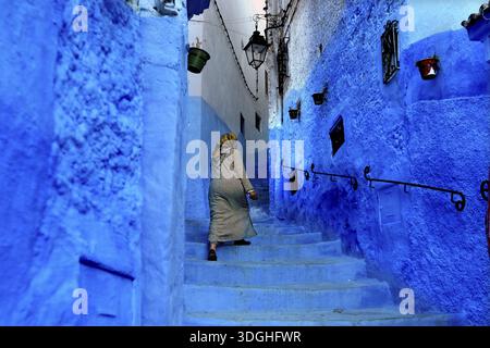 Femme en tenue traditionnelle escaladant les marches bleues d'une ruelle de la médina à Chefchaouen, Chefchaouen, Tanger-Tétouan, Maroc Banque D'Images