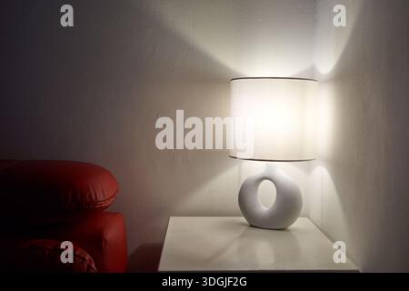 Lampe de chevet chaude à côté d'un canapé rouge, lumière douce projetant des ombres radiales sur la table blanche, base annulaire en céramique Banque D'Images