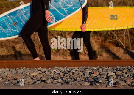 Deux surfeurs en combinaisons noires marchant le long des voies ferrées au coucher du soleil, chacun portant une planche de surf. L'un a une planche de surf bleue, l'autre jaune. Banque D'Images