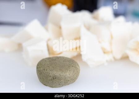 Une barre de shampooing solide ronde et texturée repose sur une surface blanche. Dans le fond flou se trouvent de gros morceaux de beurre de karité cru, un ingrédient clé pour le makin Banque D'Images