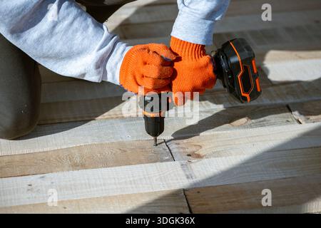 Gros plan des mains d'un ouvrier en gants orange à l'aide d'une perceuse électrique sans fil pour enfoncer une vis dans un nouveau pont en bois. Ce projet de construction DIY Banque D'Images