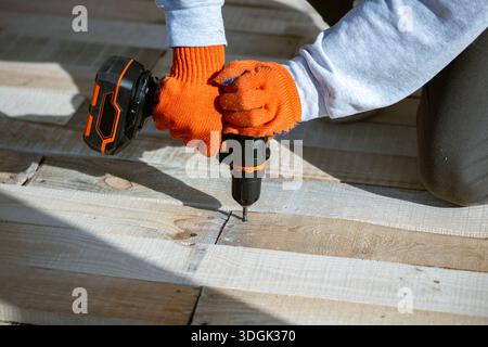 Gros plan des mains d'un ouvrier en gants orange à l'aide d'une perceuse électrique sans fil pour enfoncer une vis dans un nouveau pont en bois. Ce projet de construction DIY Banque D'Images