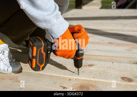Gros plan des mains d'un ouvrier en gants orange à l'aide d'une perceuse électrique sans fil pour enfoncer une vis dans un nouveau pont en bois. Ce projet de construction DIY Banque D'Images