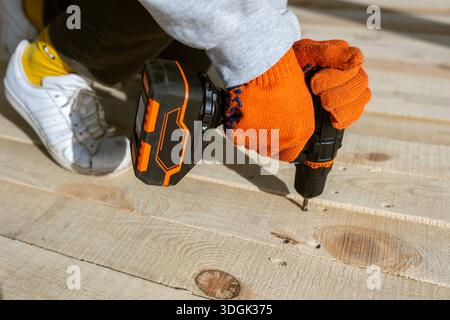 Gros plan des mains d'un ouvrier en gants orange à l'aide d'une perceuse électrique sans fil pour enfoncer une vis dans un nouveau pont en bois. Ce projet de construction DIY Banque D'Images