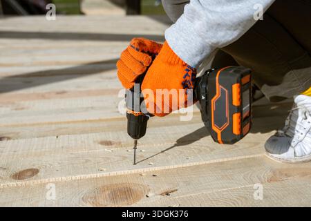 Gros plan des mains d'un ouvrier en gants orange à l'aide d'une perceuse électrique sans fil pour enfoncer une vis dans un nouveau pont en bois. Ce projet de construction DIY Banque D'Images