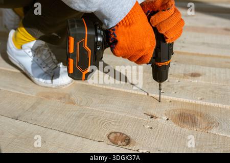 Gros plan des mains d'un ouvrier en gants orange à l'aide d'une perceuse électrique sans fil pour enfoncer une vis dans un nouveau pont en bois. Ce projet de construction DIY Banque D'Images