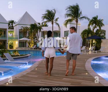 Un couple profite d'une promenade romantique le long du sentier de la piscine dans un luxueux complexe hôtelier d'Aruba. Entourés de palmiers et d'un éclairage doux, ils embra Banque D'Images