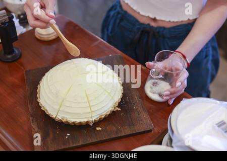 Gros plan main de femme tenant une cuillère en bois étalant la garniture de crème douce blanche sur une délicieuse tarte à la noix de coco maison sur un plateau en bois se préparant à servir Banque D'Images