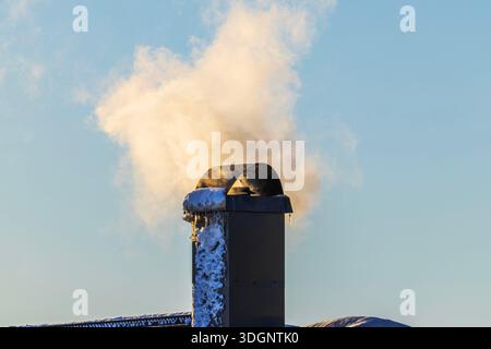 Vue rapprochée du tuyau de cheminée sur le toit de la villa avec de la fumée s'élevant dans le ciel froid d'hiver. Suède. Banque D'Images