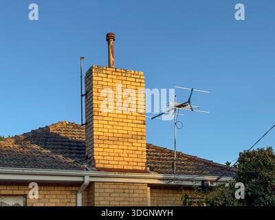 Une vue détaillée d'un toit de maison avec une cheminée en briques et une antenne, éclairée par la lumière chaude du soleil contre un ciel bleu vif. Banque D'Images