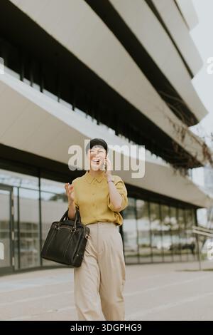 Une femme en blouse jaune et pantalon léger marche joyeusement à l'extérieur d'un bâtiment contemporain. Elle tient un sac à main noir et est engagée dans un conv. De téléphone Banque D'Images