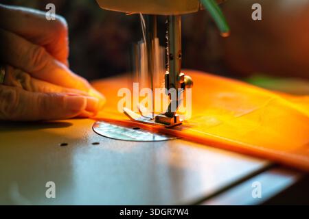 Gros plan du pied presseur de machine à coudre sur tissu orange avec une main gauche dans un cadre d'atelier lumineux et avec une faible profondeur de champ Banque D'Images