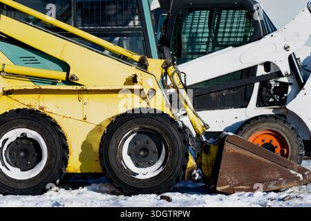 Chargeurs compacts rigides de construction stationnés côte à côte dans la neige, gros plan sur les roues jaunes du godet et la carrosserie sur les chantiers hivernaux Banque D'Images