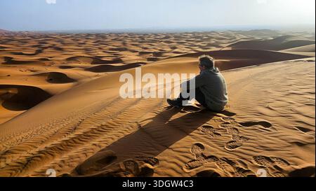 Personne solitaire dans un sweat à capuche assis sur la crête d'une dune massive de sable doré, contemplant les dunes roulantes infinies du Sahara marocain sous Banque D'Images