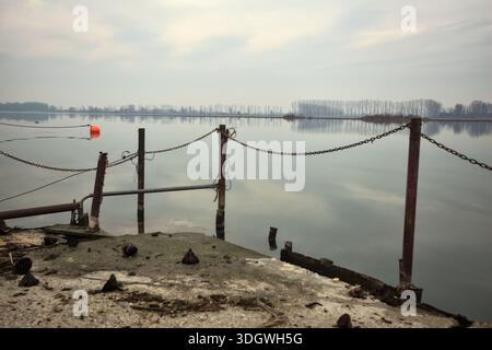 Chaîne de fer sur un coin par le bord d'une jetée au bord du lac, avec des bouées flottantes à proximité et un ciel nuageux coulé dans l'eau, dans le pays italien Banque D'Images