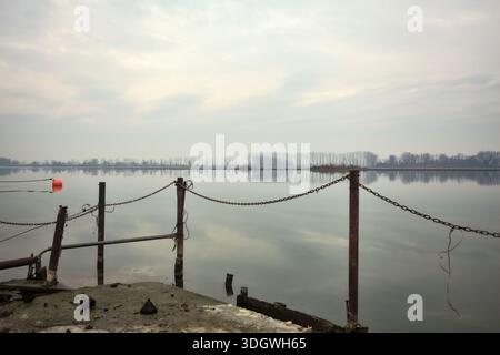 Chaîne de fer sur un coin par le bord d'une jetée au bord du lac, avec des bouées flottantes à proximité et un ciel nuageux coulé dans l'eau, dans le pays italien Banque D'Images