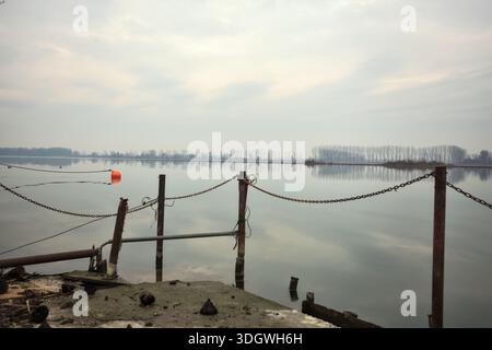 Chaîne de fer sur un coin par le bord d'une jetée au bord du lac, avec des bouées flottantes à proximité et un ciel nuageux coulé dans l'eau, dans le pays italien Banque D'Images