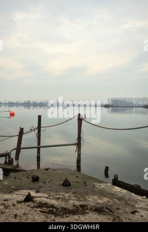 Chaîne de fer sur un coin par le bord d'une jetée au bord du lac, avec des bouées flottantes à proximité et un ciel nuageux coulé dans l'eau, dans le pays italien Banque D'Images