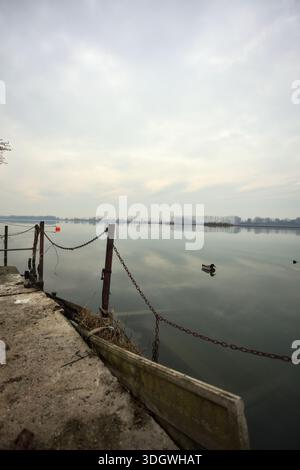 Chaîne de fer sur un coin par le bord d'une jetée au bord du lac, avec des bouées flottantes à proximité et un ciel nuageux coulé dans l'eau, dans le pays italien Banque D'Images