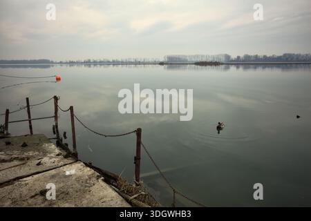 Chaîne de fer sur un coin par le bord d'une jetée au bord du lac, avec des bouées flottantes à proximité et un ciel nuageux coulé dans l'eau, dans le pays italien Banque D'Images